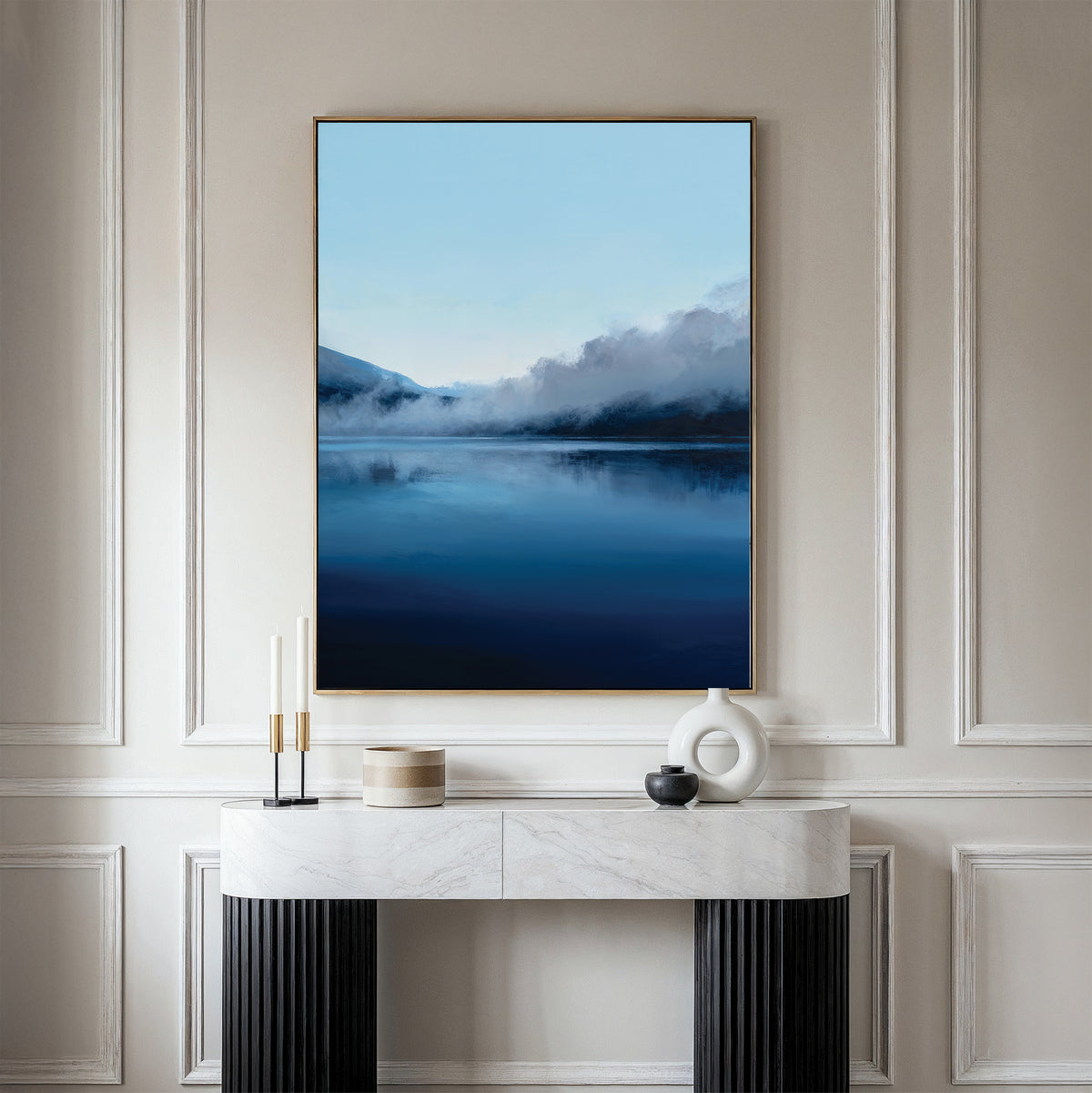 A modern living space with a white marble console table, black legs, and minimalist Nordic decor, featuring the Fjord Mist Canvas Art - Vertical—a large serene landscape of a blue lake with misty mountains above the console.