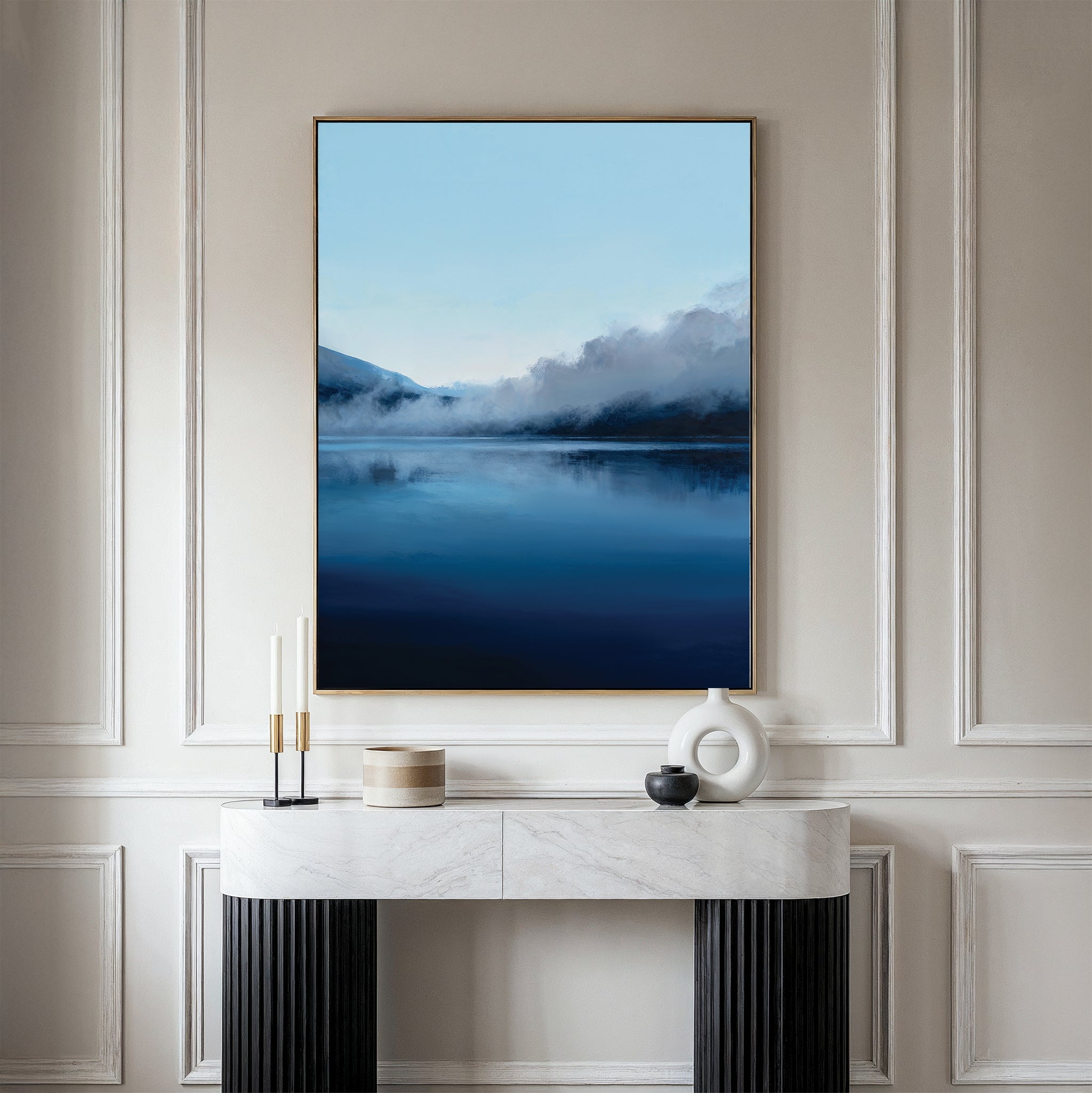 A modern living space with a white marble console table, black legs, and minimalist Nordic decor, featuring the Fjord Mist Canvas Art - Vertical—a large serene landscape of a blue lake with misty mountains above the console.
