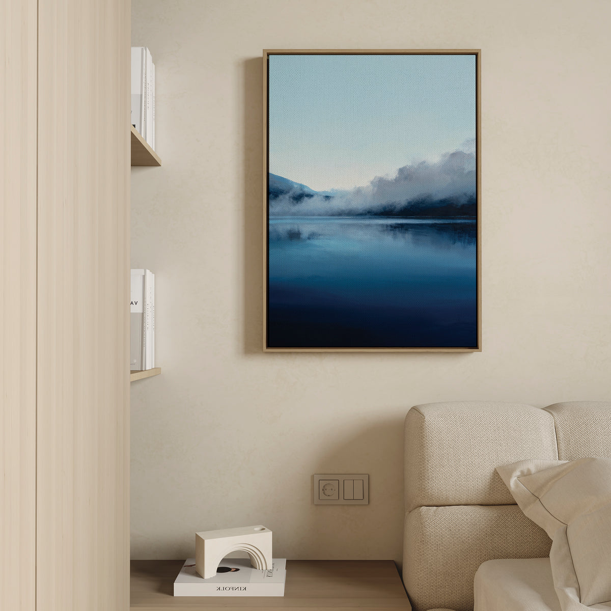 The Fjord Mist Canvas Art - Vertical, featuring a serene misty blue lake and mountains, hangs on a beige wall above a light-colored sofa; nearby, books and a geometric decor piece on a shelf enhance the Nordic vibe.