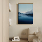 The Fjord Mist Canvas Art - Vertical, featuring a serene misty blue lake and mountains, hangs on a beige wall above a light-colored sofa; nearby, books and a geometric decor piece on a shelf enhance the Nordic vibe.