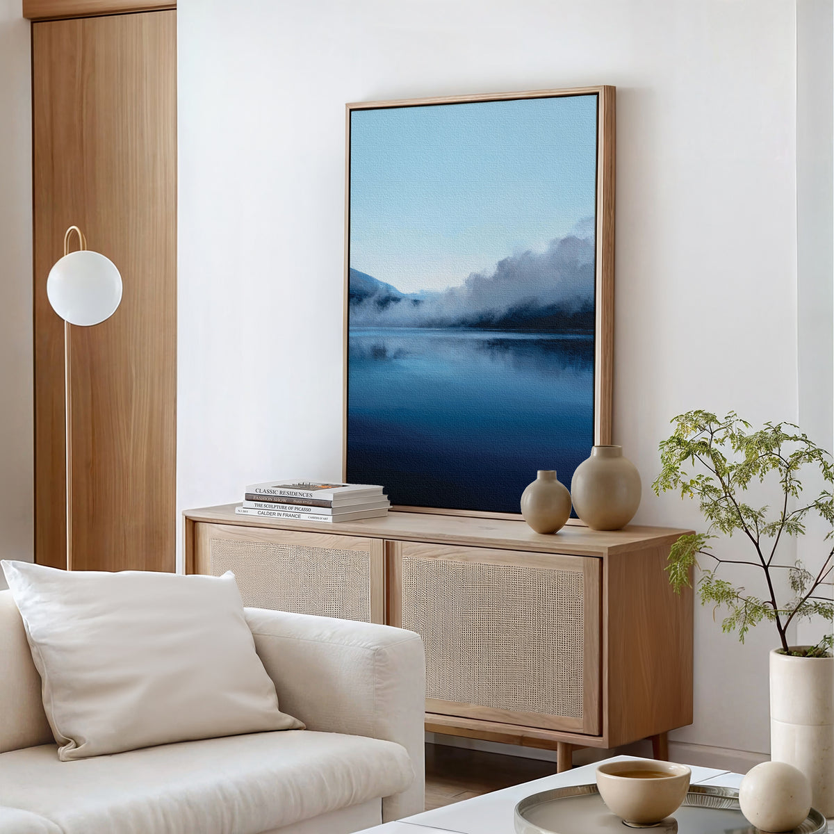 A modern living room with neutral tones showcases the Fjord Mist Canvas Art - Vertical of a misty lake, ceramic vases, stacked books on a wooden sideboard, a white cushioned sofa, round hanging lamp, and potted plant for timeless Nordic decor.