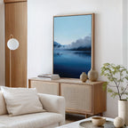 A modern living room with neutral tones showcases the Fjord Mist Canvas Art - Vertical of a misty lake, ceramic vases, stacked books on a wooden sideboard, a white cushioned sofa, round hanging lamp, and potted plant for timeless Nordic decor.