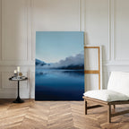 A minimalist room with herringbone floors, a white chair, and Nordic decor features a round black side table with candles and the Fjord Mist Canvas Art - Vertical propped against a white paneled wall.