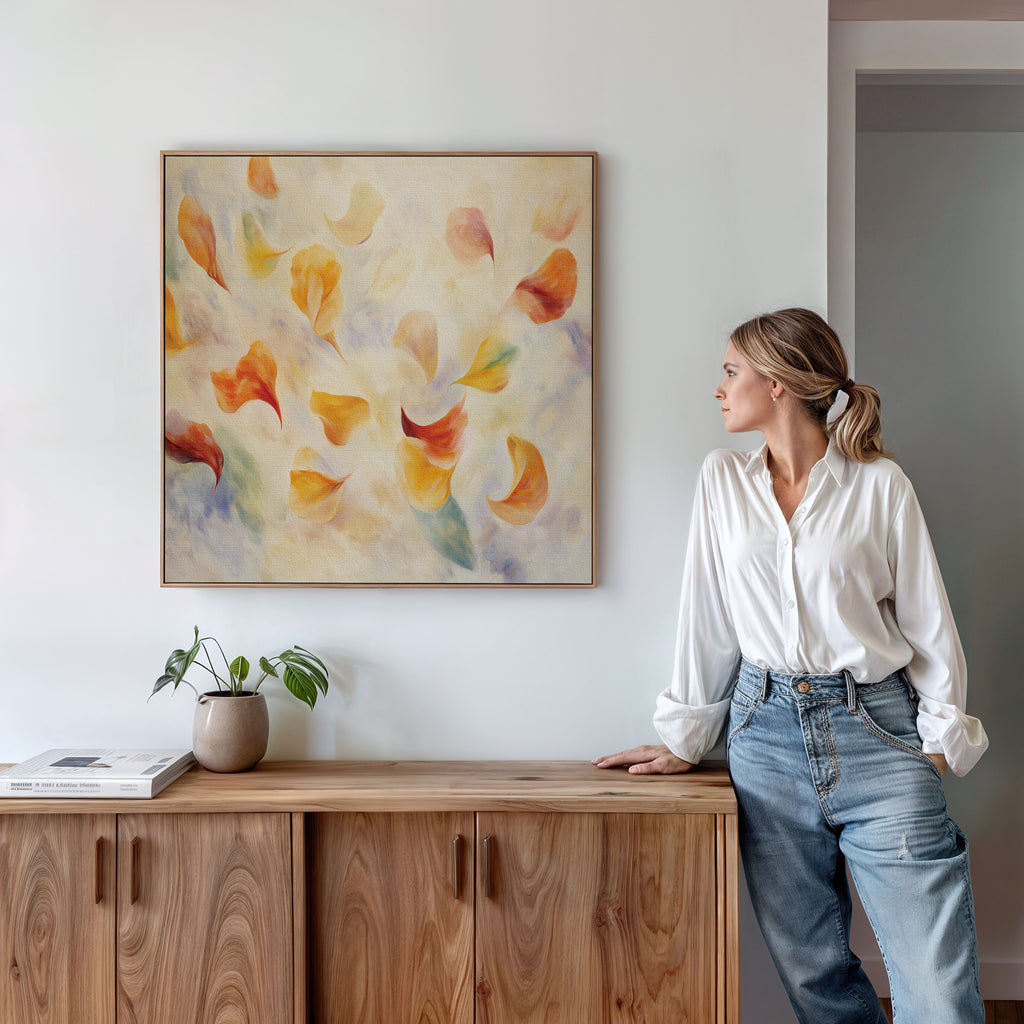 A woman in a white blouse and jeans leans against a wooden cabinet, admiring the Floating Petals Canvas Art - Square, which features warm-toned swirling shapes on a pale background. A potted plant and book add natural elegance to the setting.