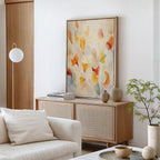A modern living room with a neutral sofa, wooden sideboard, ceramics, and a potted plant features “Floating Petals Canvas Art - Vertical,” adding organic elegance in soft orange, yellow, and red tones to the wall.
