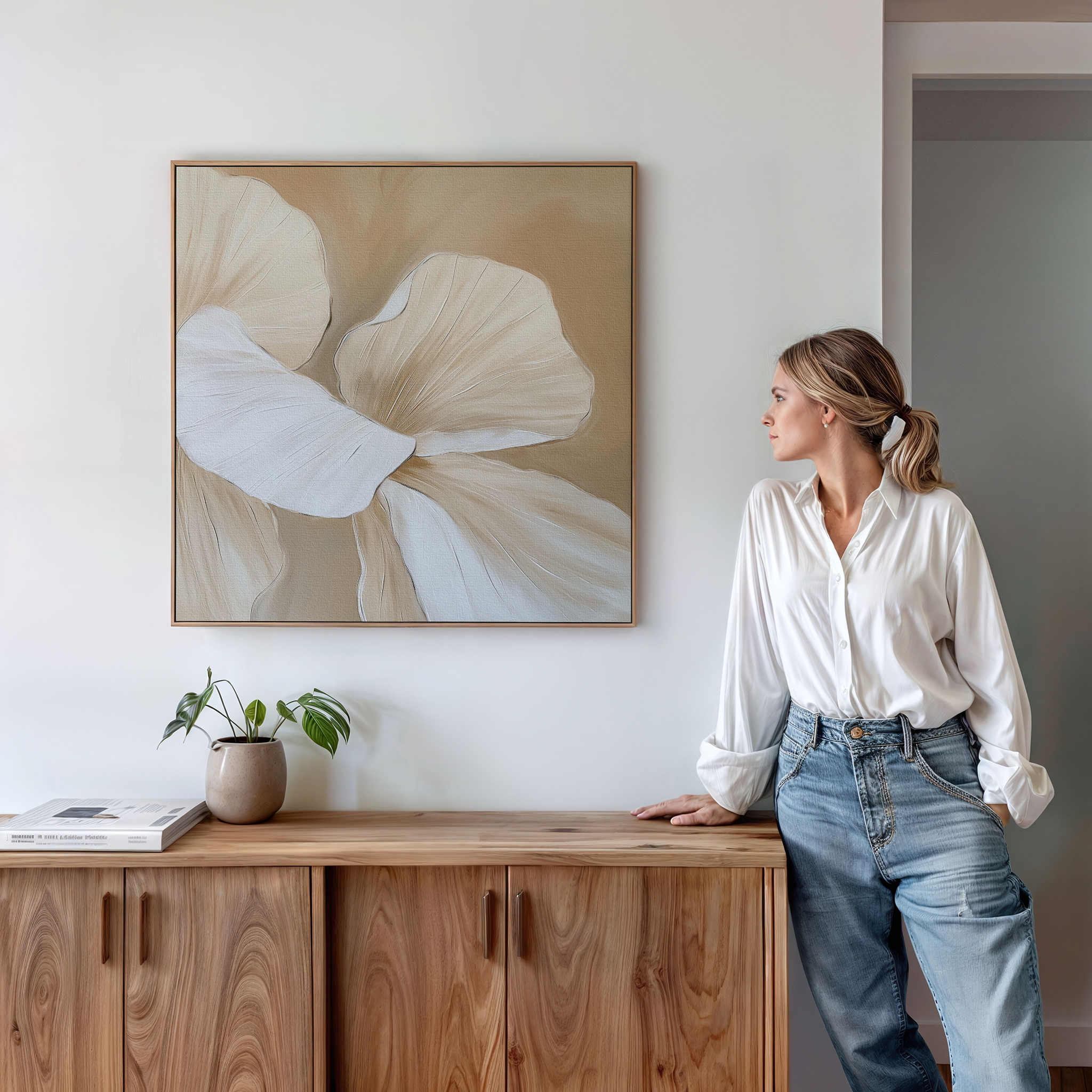 A woman in a white blouse and jeans stands by a wooden sideboard decorated with a potted plant and books, admiring the Floral Whisper Abstract Canvas Art—a large beige and white abstract print—hanging on the white wall.