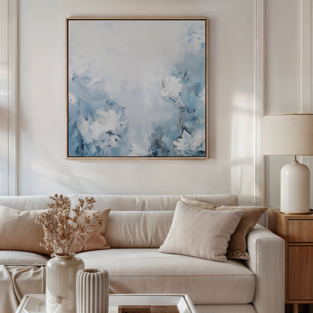 A modern living room features a beige sofa with neutral pillows, a side table, a lamp, dried flowers in a vase, and captivating wall art: the Spring Hues Abstract Canvas Art in blue and white. Soft natural light illuminates the space.