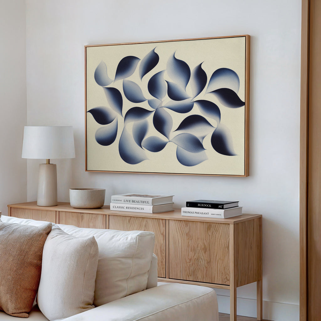 A modern living room with a light wood sideboard, books, a lamp, and ceramic bowls. Above the sideboard hangs the Flowing Shadows Canvas Art - Horizontal, displaying blue and gray organic shapes on a beige background.