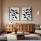 A modern living room features a wooden sideboard, beige sofa, matching coffee table, and "Flowing Shadows - Set of 2" abstract canvas prints above the sideboard. A small plant and books complete the cozy decor.