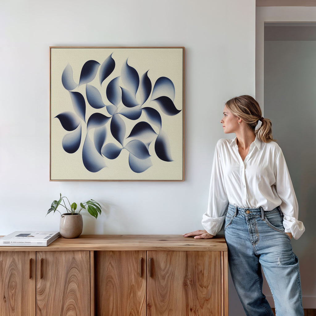 A woman in a white blouse and jeans stands beside a wooden cabinet, admiring the Flowing Shadows Canvas Art - Square. A small potted plant and books decorate the modern living room, enhancing the stylish atmosphere.