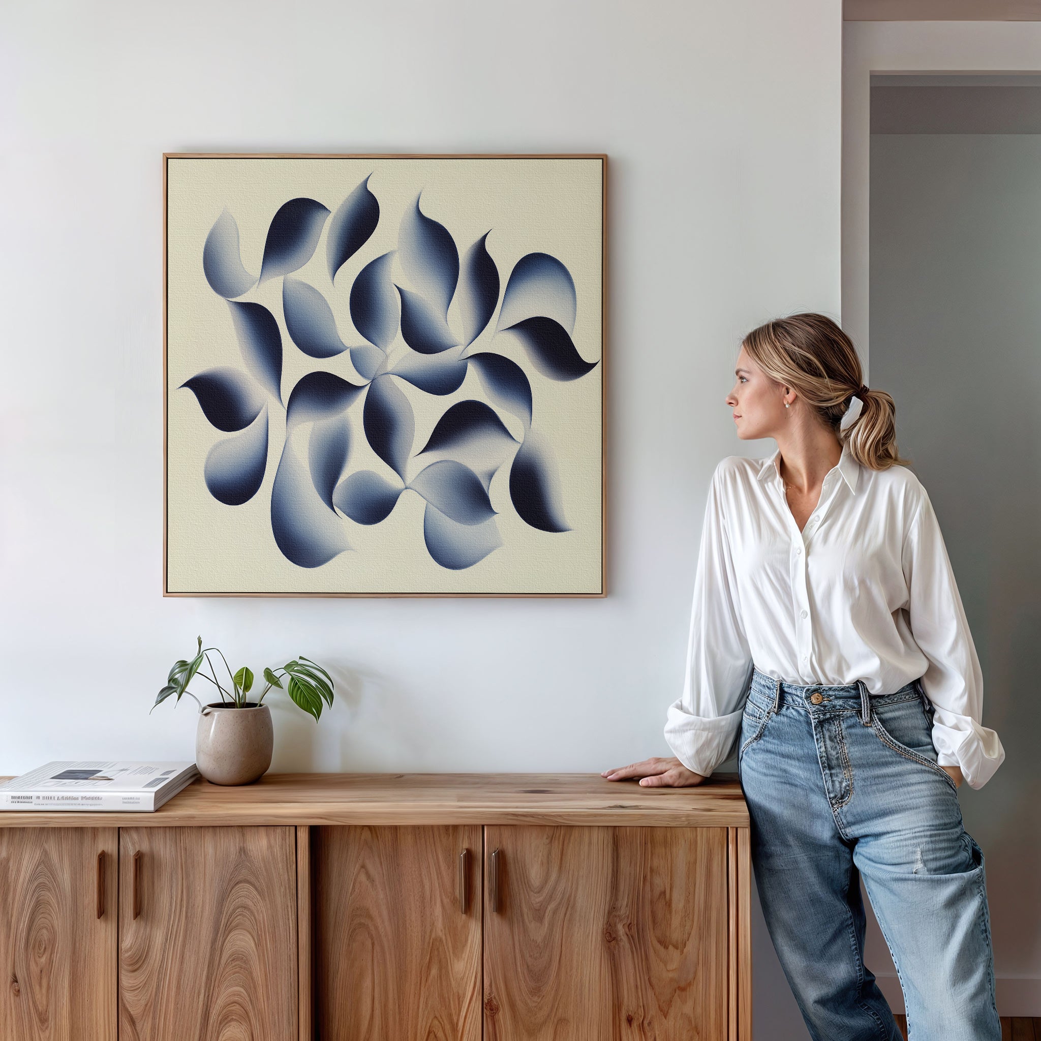 A woman in a white blouse and jeans stands beside a wooden cabinet, admiring the Flowing Shadows Canvas Art - Square. A small potted plant and books decorate the modern living room, enhancing the stylish atmosphere.