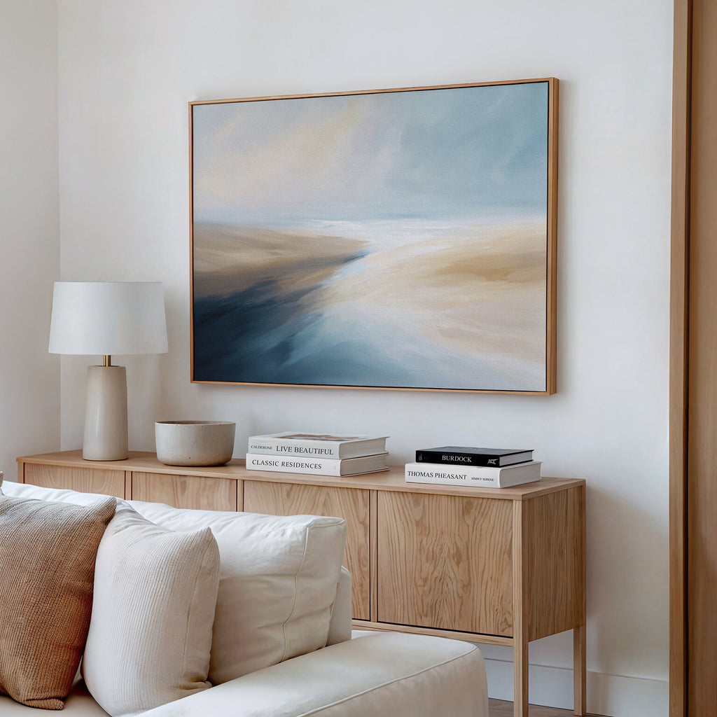 A modern living room featuring the Fluid Motion Canvas Art - Horizontal in soft blue, beige, and white tones pairs with a wooden sideboard, stacked books, a ceramic bowl, and a white lamp for a serene coastal vibe.