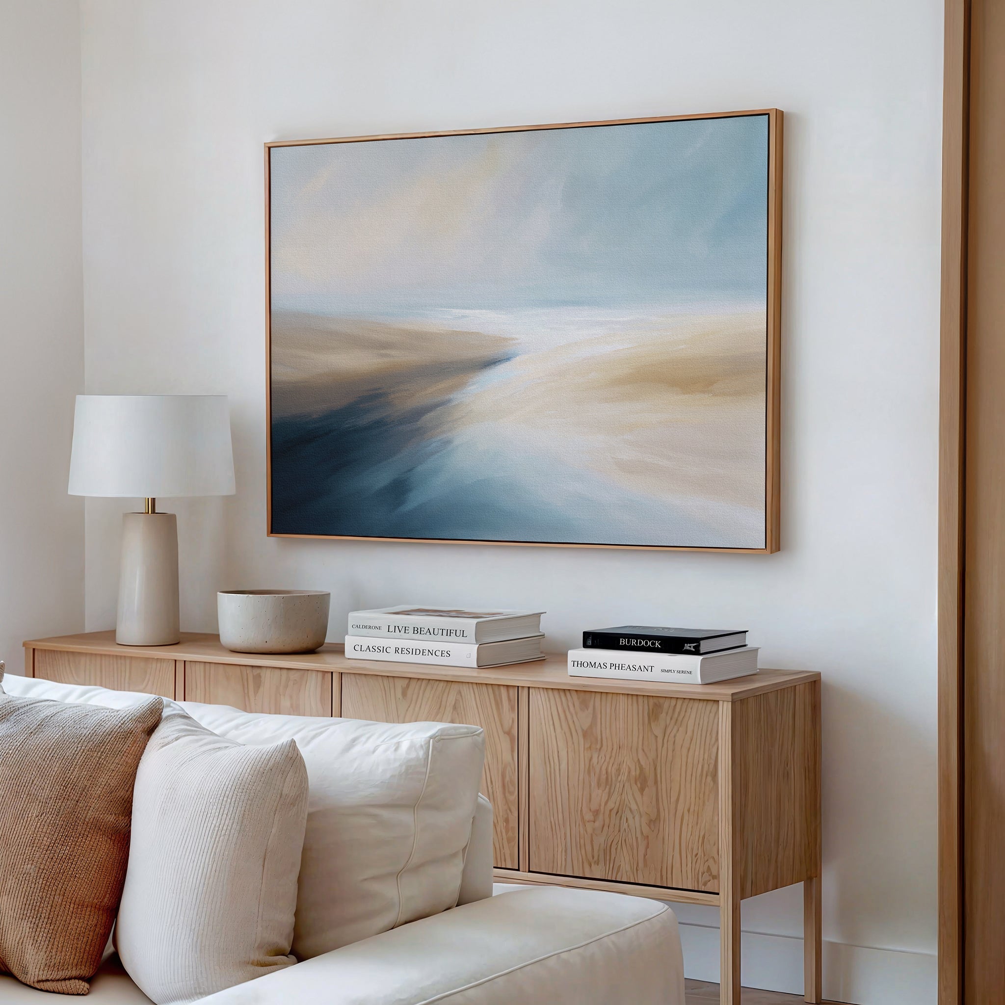 A modern living room featuring the Fluid Motion Canvas Art - Horizontal in soft blue, beige, and white tones pairs with a wooden sideboard, stacked books, a ceramic bowl, and a white lamp for a serene coastal vibe.