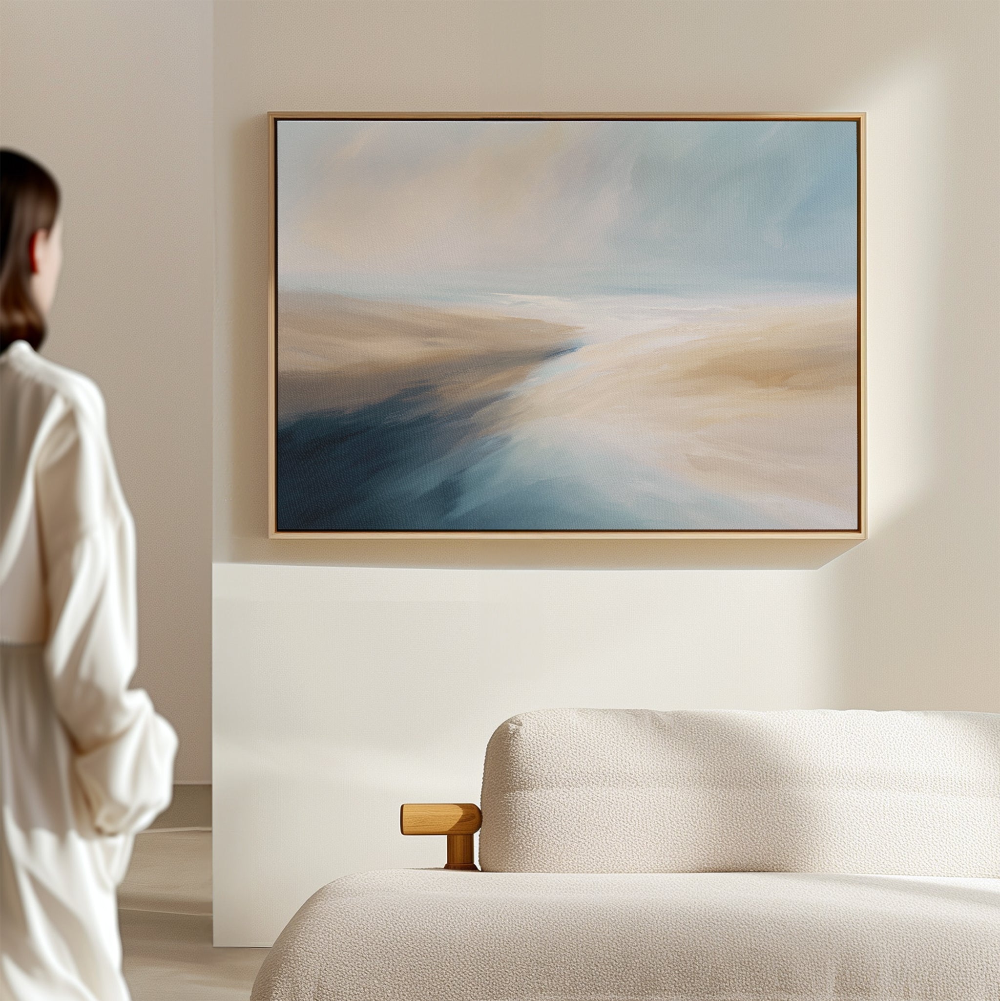 A person in a white shirt stands in a bright, minimal room, admiring the Fluid Motion Canvas Art - Horizontal with blue and beige tones above a cream sofa—evoking serene coastal vibes ideal for modern living rooms.