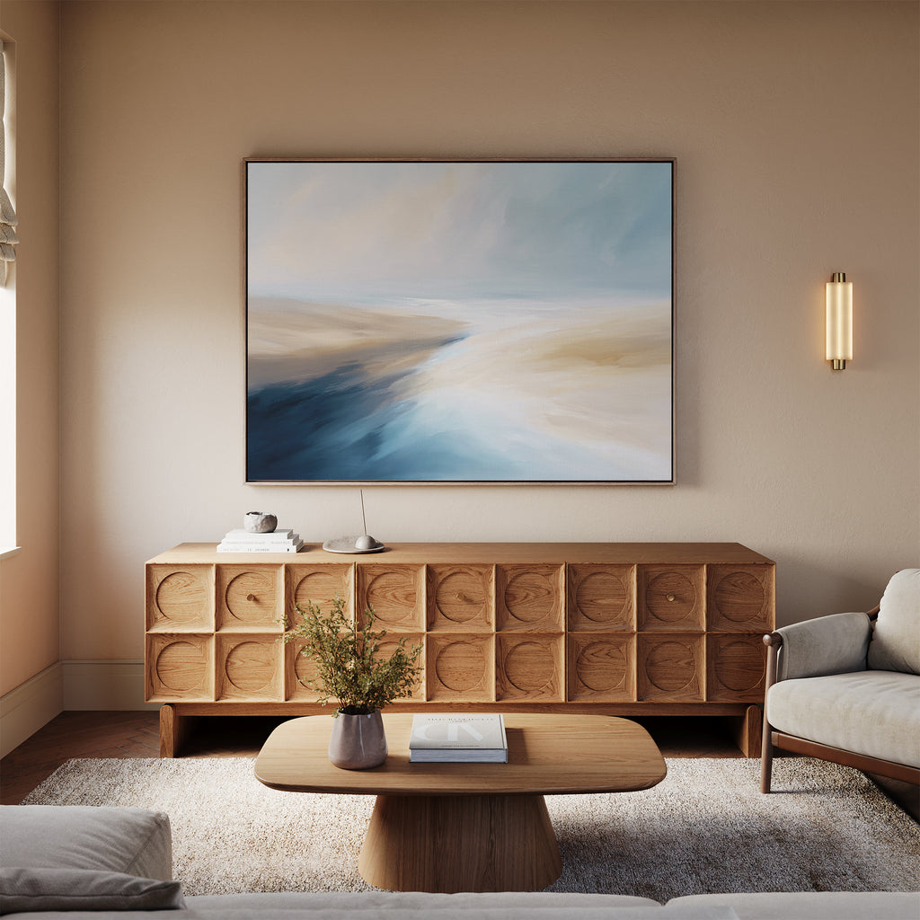 A modern living room features a wooden coffee table, light rug, and beige sofa for a coastal vibe. Above the sideboard hangs the Fluid Motion Canvas Art - Horizontal in soft blue and beige, while a small plant decorates the table.
