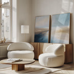 A modern living room with two curved white armchairs, a low wooden coffee table, a floor lamp, and Fluid Motion - Set Of 2 coastal wall art in blue and beige on a wooden sideboard by a large window.
