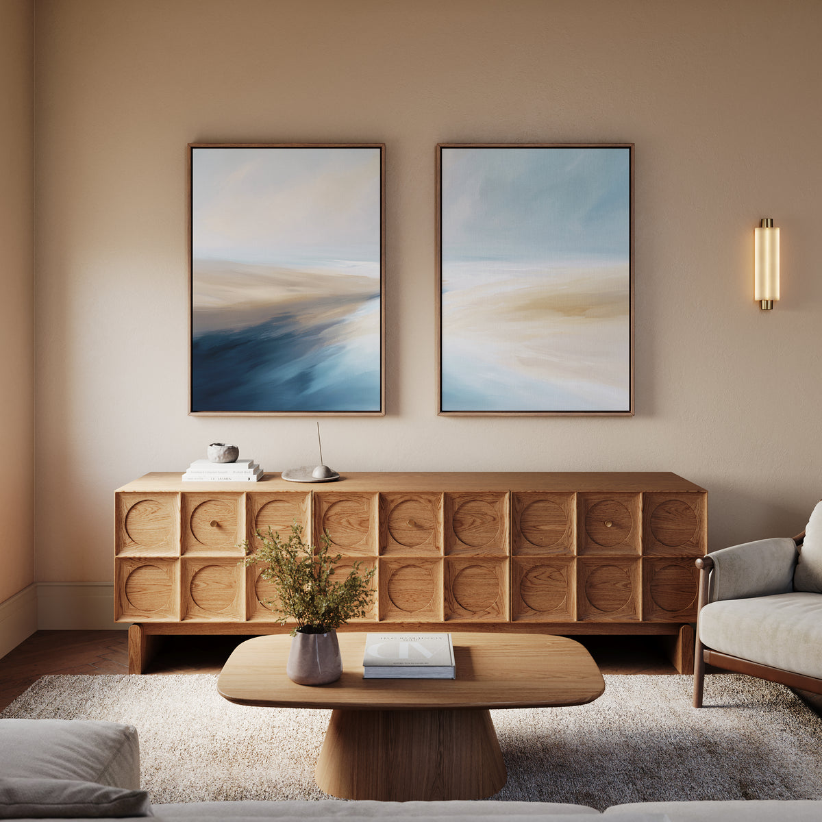 A modern living room featuring the Fluid Motion - Set Of 2 abstract blue and beige coastal wall art, a wooden coffee table, sideboard, neutral armchairs, and a small plant. Soft lighting adds coziness—perfect for stylish modern decor.