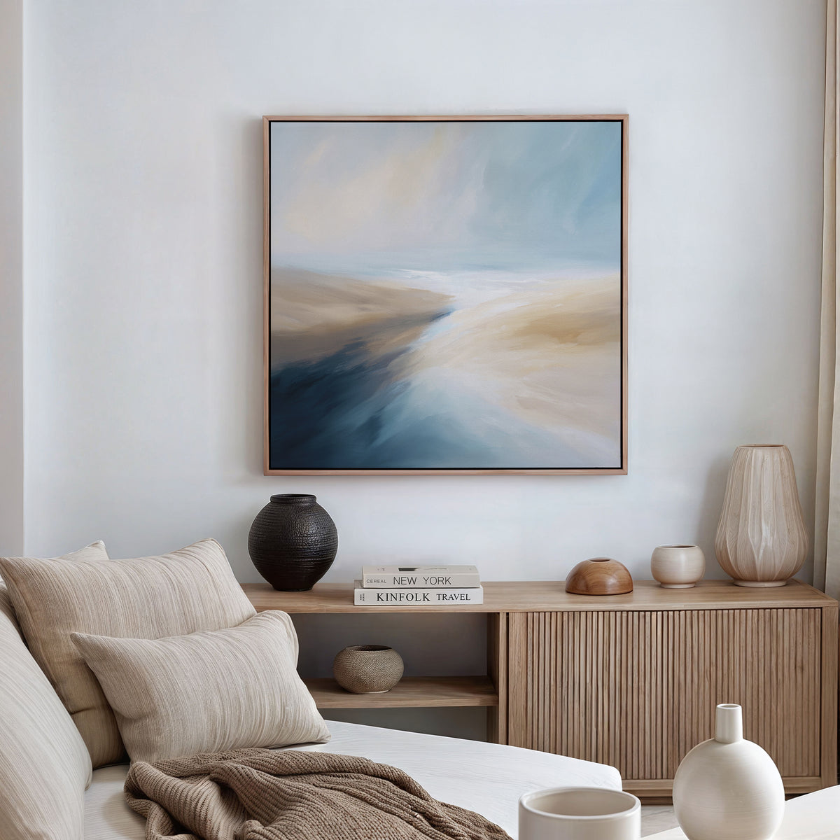 A modern living room features a beige sofa with neutral cushions, a wooden sideboard displaying vases and decor, and the Fluid Motion Canvas Art - Square in blue and beige tones above, creating a subtle coastal vibe.