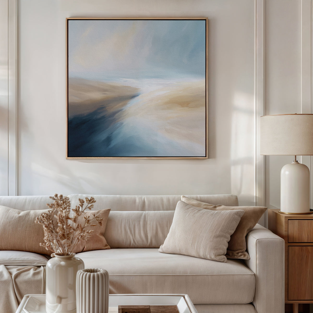 A modern living room with a beige sofa, neutral cushions, a wooden side table with a cream lamp, dried flowers in a vase, and the Fluid Motion Canvas Art - Square above the couch creates a relaxed coastal atmosphere.