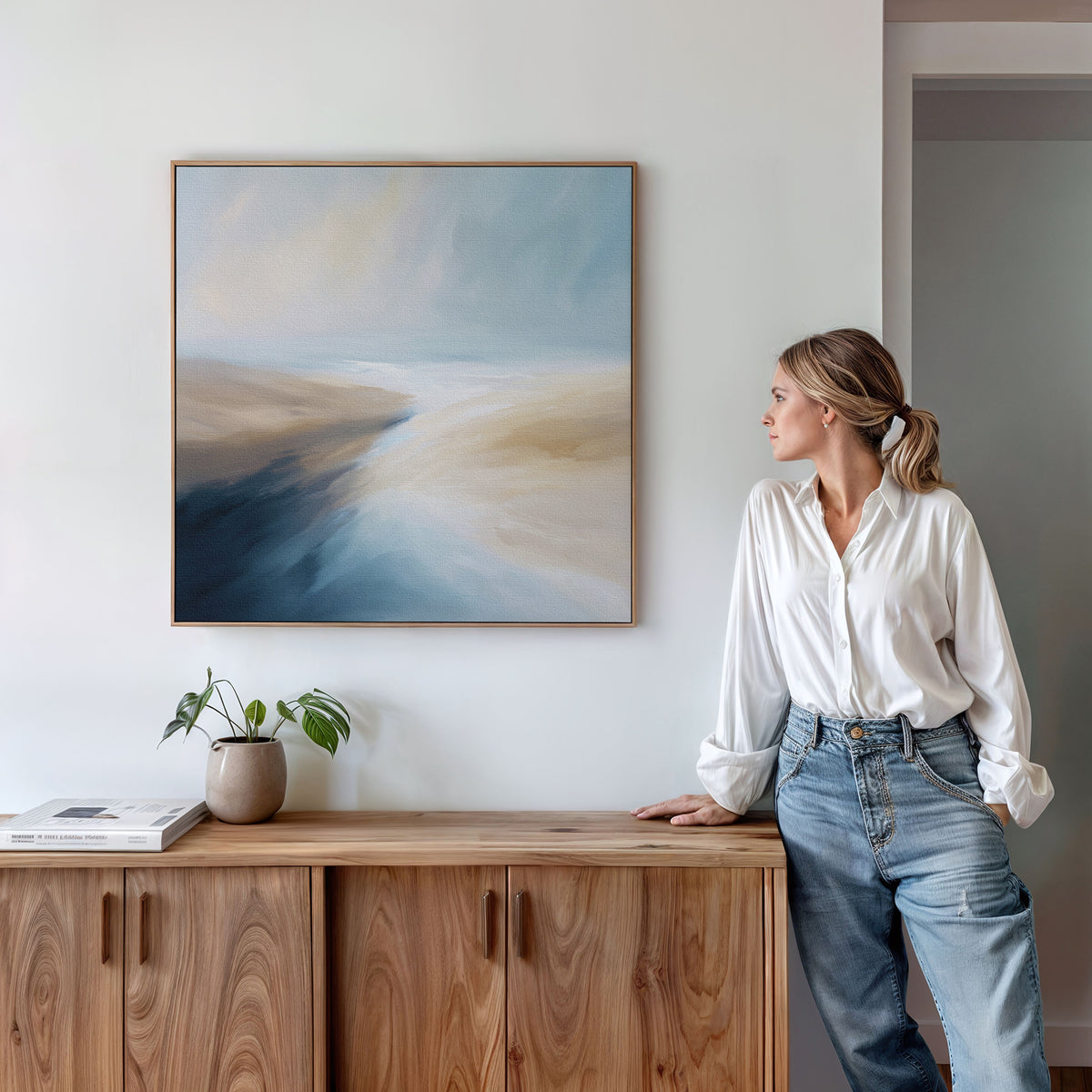 A woman in a white shirt and jeans leans against a wooden cabinet, admiring the Fluid Motion Canvas Art - Square with its soft blue, beige, and white tones. A plant and books on the cabinet enhance the coastal vibe of modern living rooms.