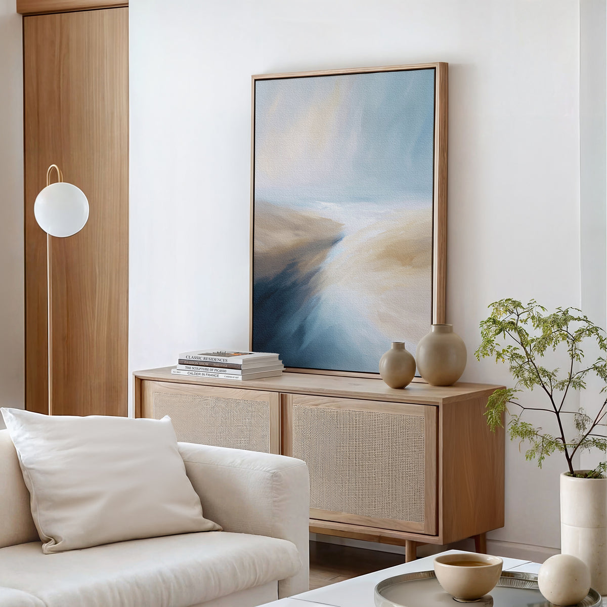 A modern living room features the Fluid Motion Canvas Art - Vertical in blue and beige tones above a light wood sideboard with vases and books. A white couch and potted plant complete the minimalist decor.