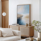 A modern living room features the Fluid Motion Canvas Art - Vertical in blue and beige tones above a light wood sideboard with vases and books. A white couch and potted plant complete the minimalist decor.