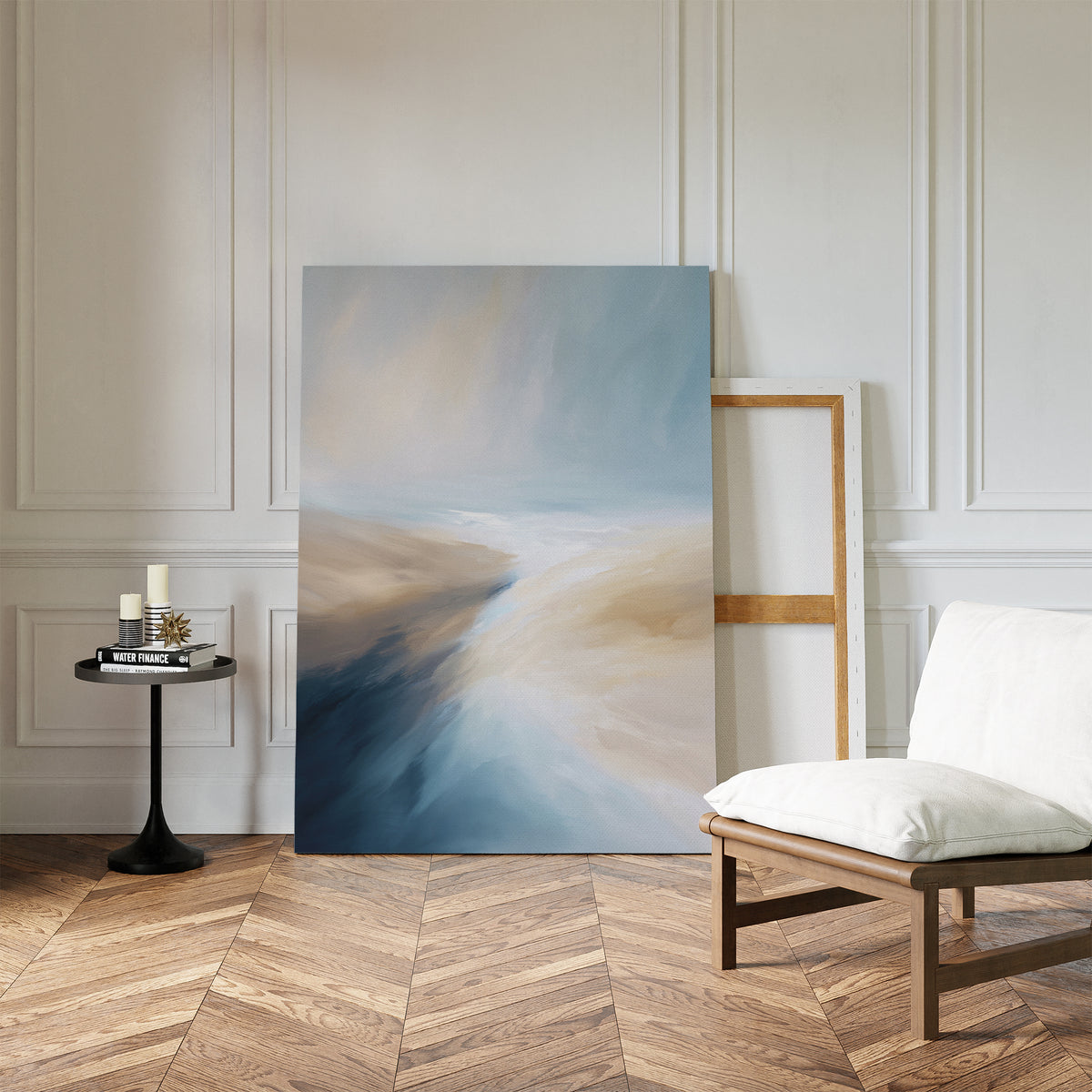 A modern, minimalist room features the Fluid Motion Canvas Art - Vertical—an abstract piece in blue and beige—leaning against a white paneled wall beside an empty frame, a cushioned chair, and a black side table with candles and books.
