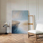 A modern, minimalist room features the Fluid Motion Canvas Art - Vertical—an abstract piece in blue and beige—leaning against a white paneled wall beside an empty frame, a cushioned chair, and a black side table with candles and books.