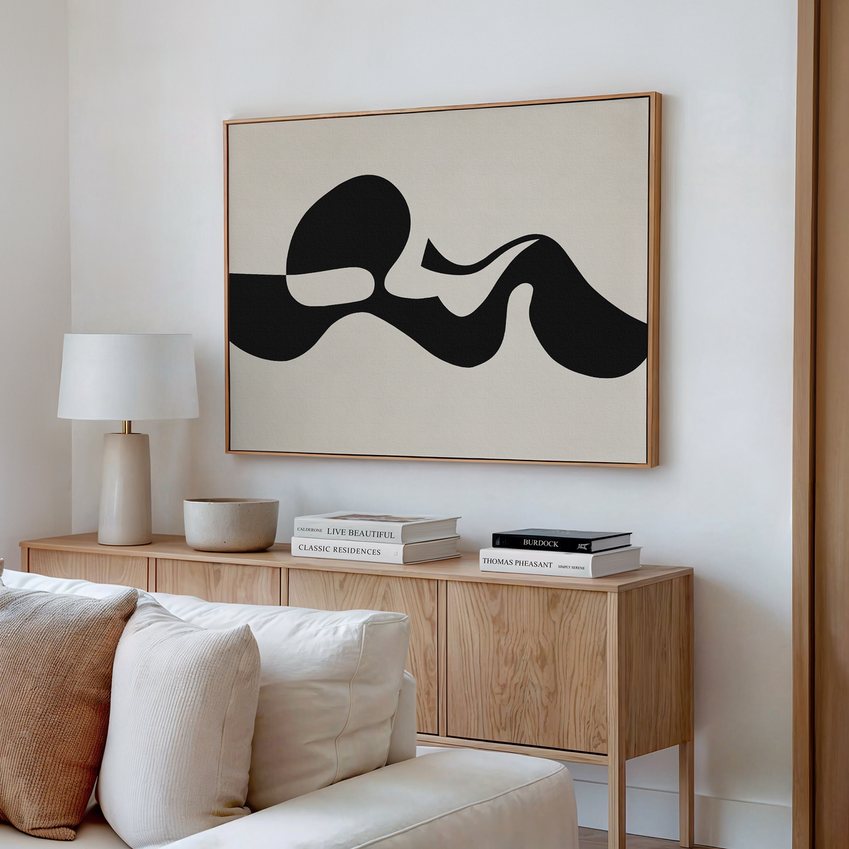 A modern living room features a light wood sideboard, white lamp, stacked books, and the Fluid Forms Abstract Canvas Art with bold black shapes in a neutral palette mounted above a white couch.