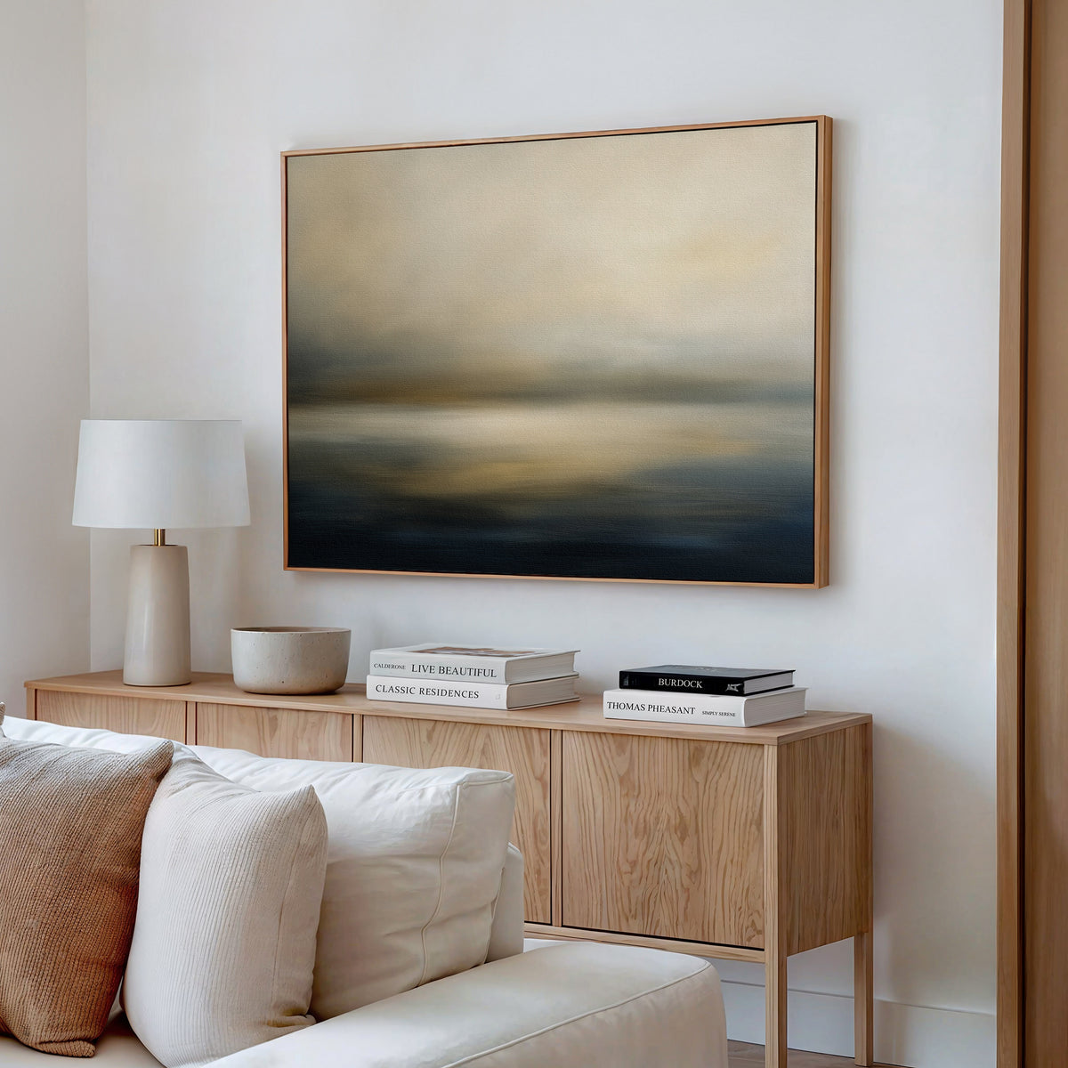 A modern living room features a wooden sideboard with books, a lamp, and a bowl. Above it hangs the Foggy Bay Impressions Canvas Art - Horizontal in soft beige and blue tones. A white sofa is placed in the foreground.
