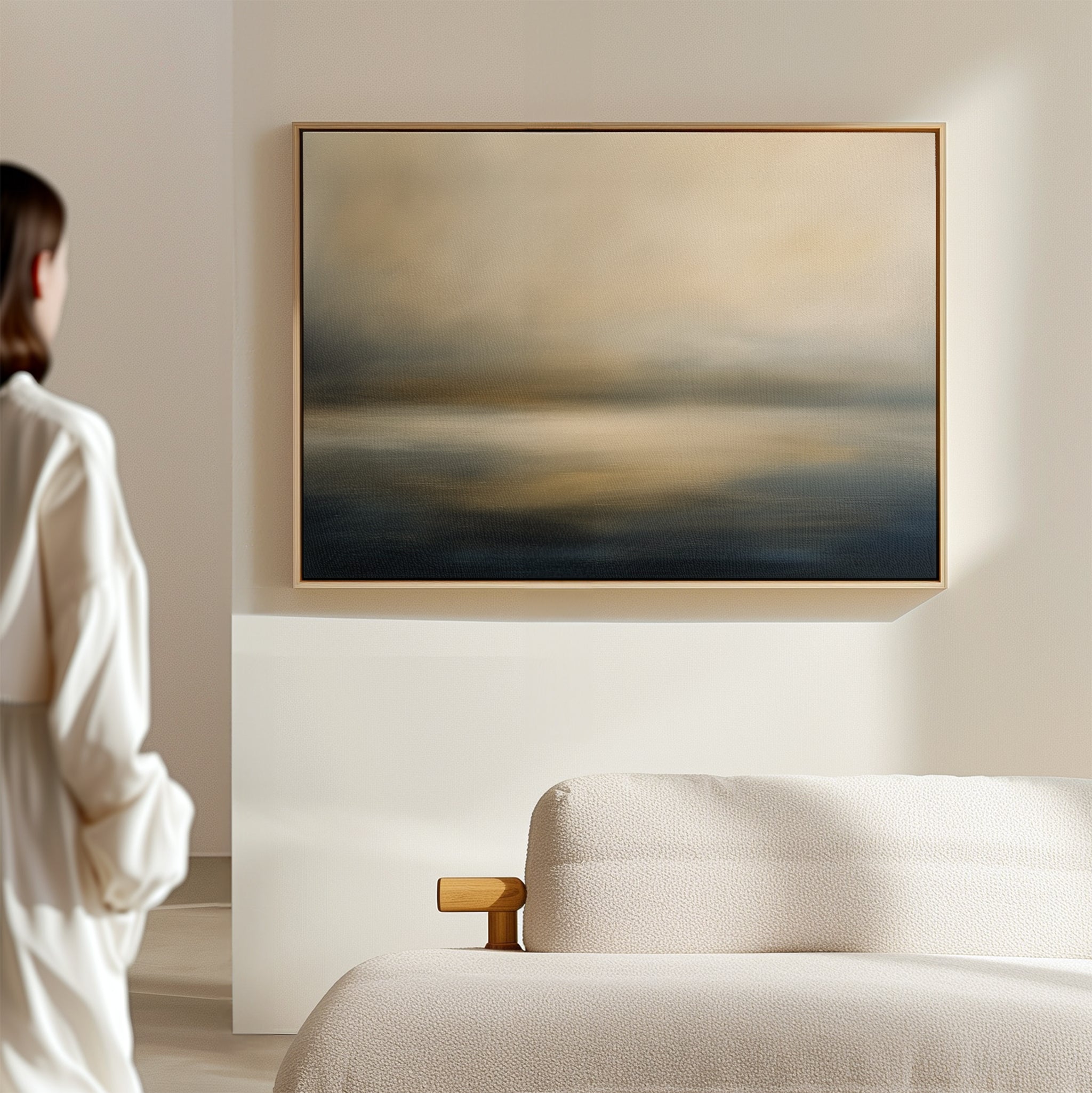 A person stands near a cream-colored sofa, admiring the Foggy Bay Impressions Canvas Art—horizontal abstract wall art in soft blue, beige, and gold tones that enhances the serene Nordic Park-inspired decor.