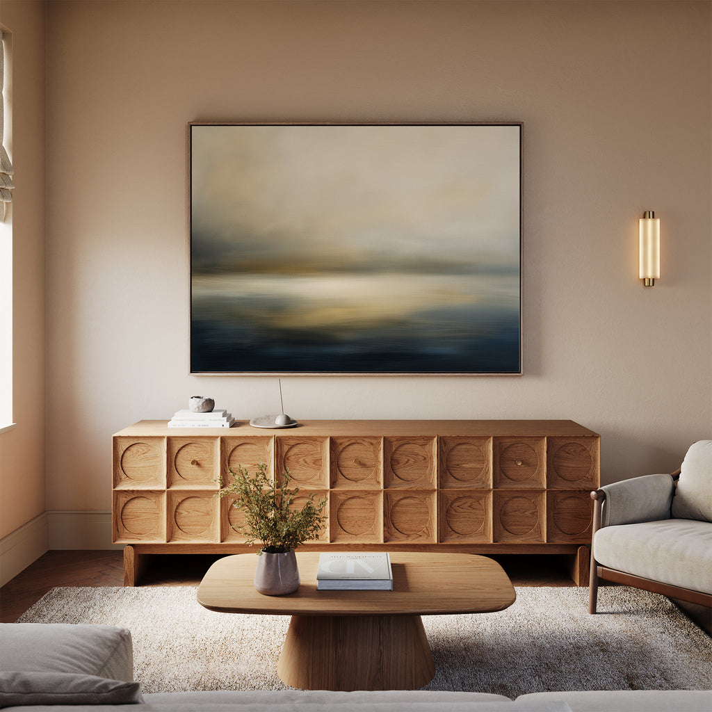 A modern living room with beige walls, the Foggy Bay Impressions Canvas Art - Horizontal above a wooden sideboard, a round coffee table with flowers, and cream sofas on a light rug creates serene Nordic Park-inspired décor.