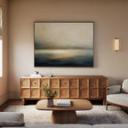 A modern living room with beige walls, the Foggy Bay Impressions Canvas Art - Horizontal above a wooden sideboard, a round coffee table with flowers, and cream sofas on a light rug creates serene Nordic Park-inspired décor.