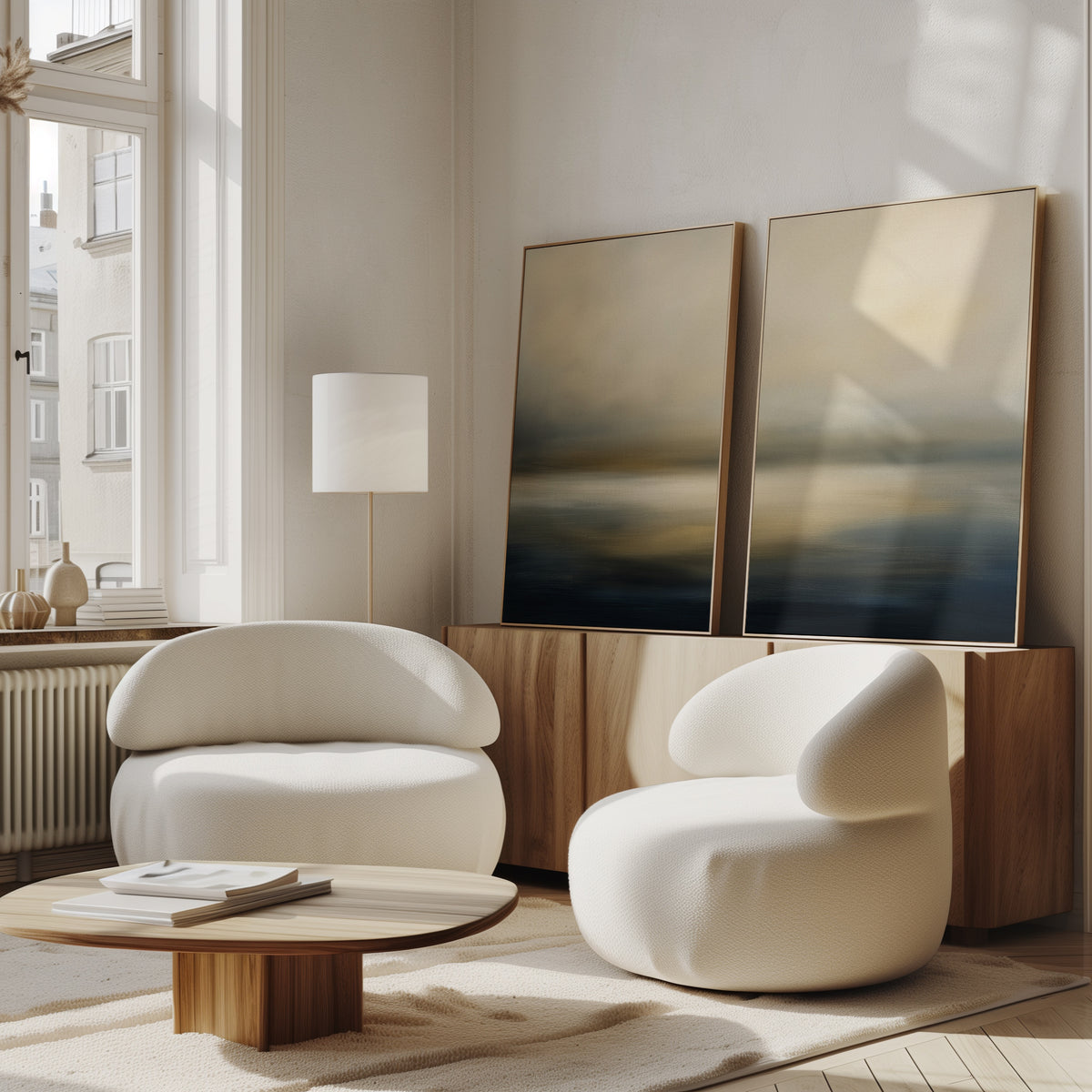 A modern living room features two white curved armchairs, a round wooden coffee table, a wooden sideboard, and the Foggy Bay Impressions - Set Of 2 abstract art resting against the wall. Soft natural light streams in through tall windows.