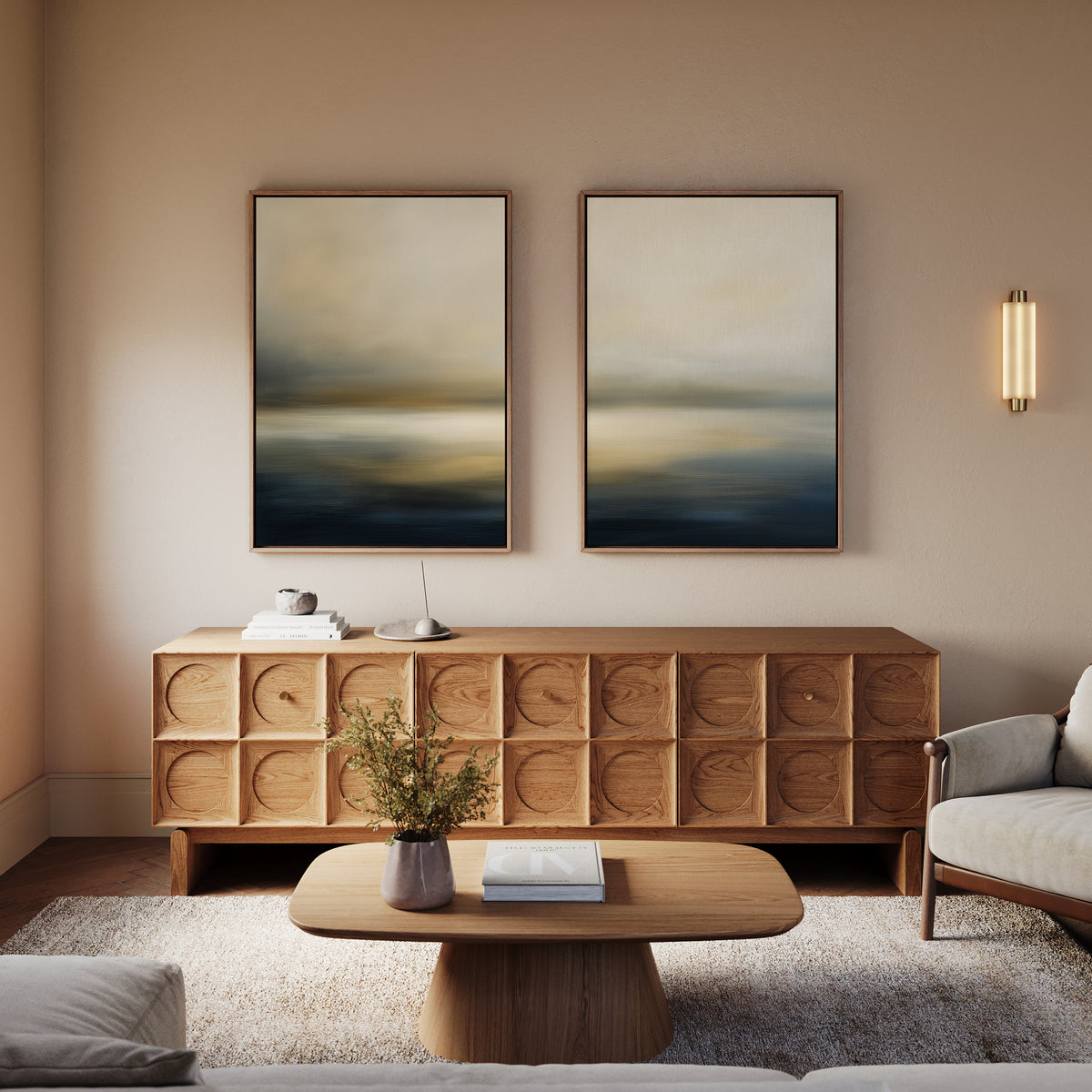 A modern living room features a wooden sideboard, coffee table, light sofa, and Foggy Bay Impressions - Set Of 2 wall art. A wall sconce adds warmth, while a vase with greenery sits on the coffee table.