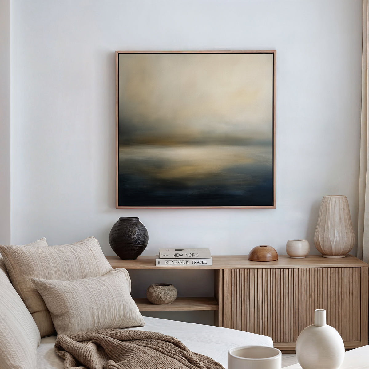 A modern living room features a beige sofa with striped pillows, a wooden sideboard holding vases and books, and the Foggy Bay Impressions Canvas Art - Square—a serene abstract painting in soft blue, beige, and gray tones.