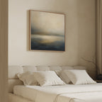 A minimalist bedroom features a beige upholstered bed with white bedding and the Foggy Bay Impressions Canvas Art - Square in soft blue, gray, and beige tones above the headboard. A bare branch in a vase sits on the nightstand.