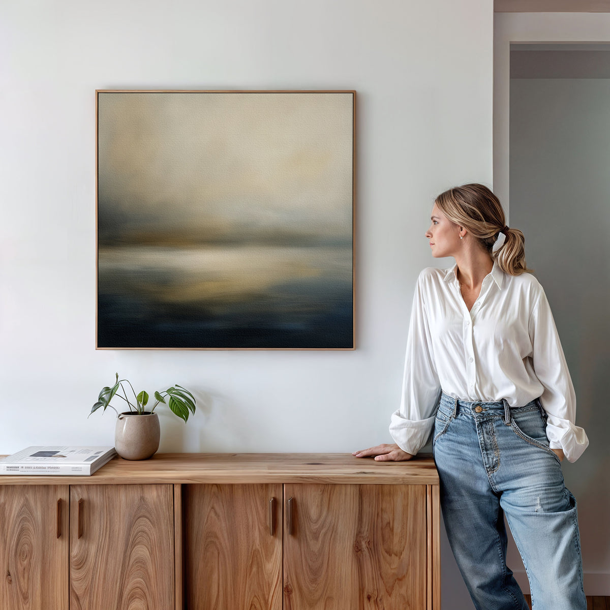 A woman in a white blouse and jeans leans against a wooden cabinet, gazing at the Foggy Bay Impressions Canvas Art - Square above, with a small plant and closed book resting on the cabinet.