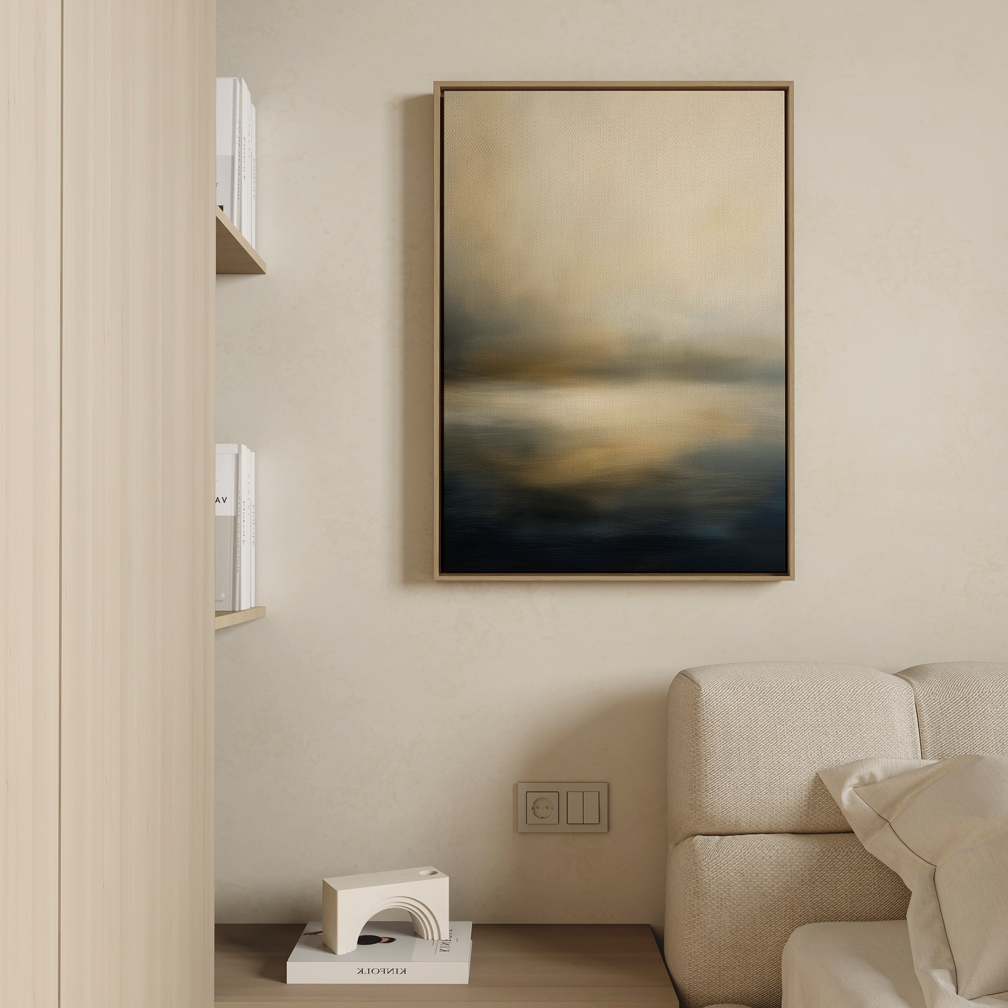 A minimal living room corner features a beige textured sofa, a wooden shelf with books and a sculpture, and the Foggy Bay Impressions Canvas Art - Vertical, adding serene elegance with its soft beige, gold, and dark blue tones.