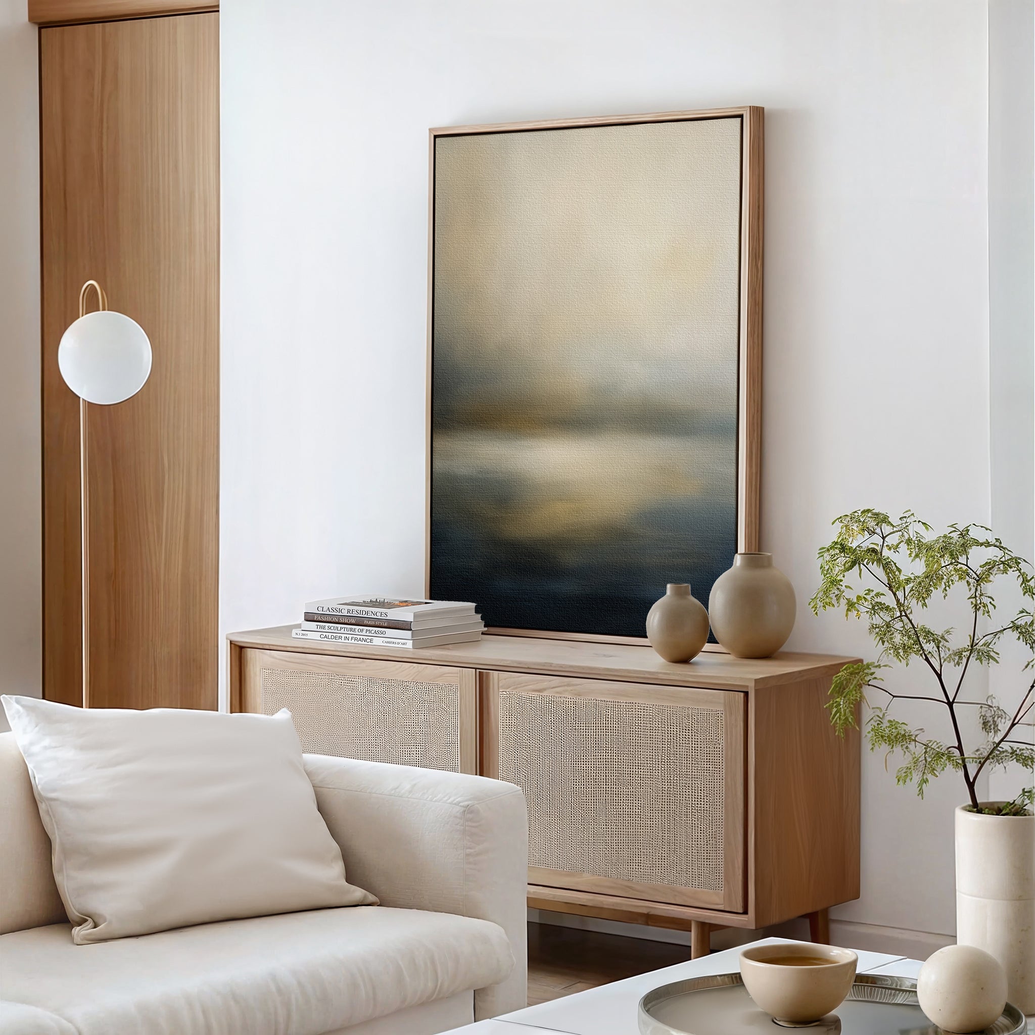 A modern living room with a light wood sideboard, Foggy Bay Impressions Canvas Art - Vertical, neutral vases, stacked books, a round pendant light, a white sofa with cushions, and a leafy plant in a beige pot exudes serene elegance.