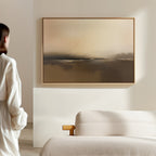 A person stands in a minimal, light-filled room with a beige sofa and the "Foggy Beach Expanse Canvas Art - Horizontal" hanging on the wall.