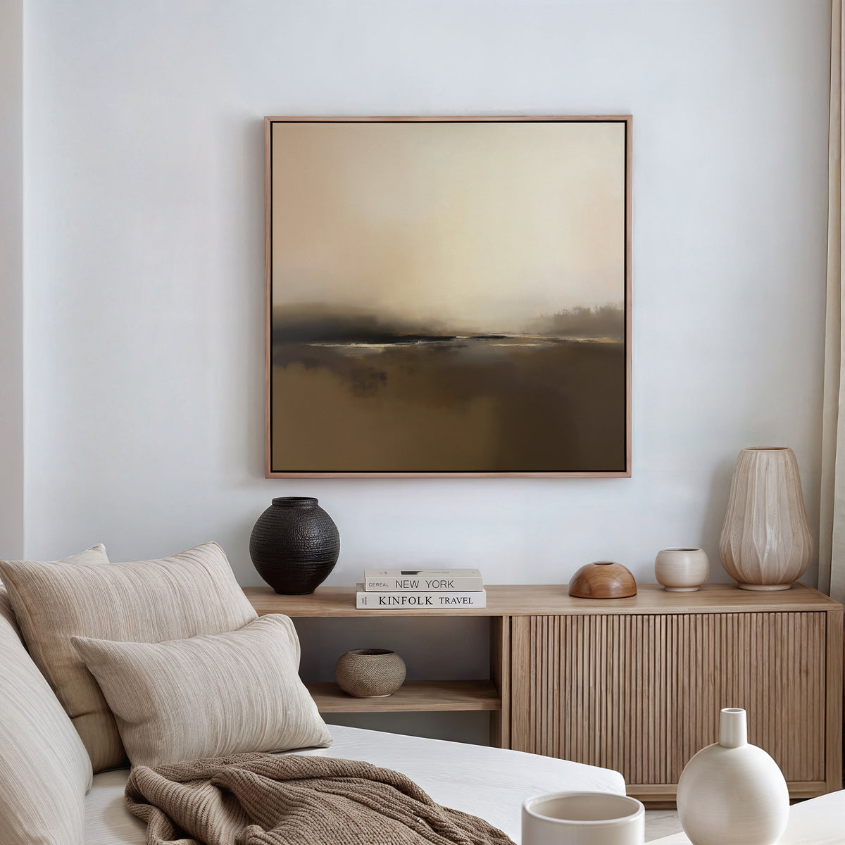 A minimal, modern living room with a neutral palette features a beige sofa, striped pillows, a wooden sideboard with vases and books, and Nordic decor touches like the Foggy Beach Expanse Canvas Art - Square in soft brown and cream tones.