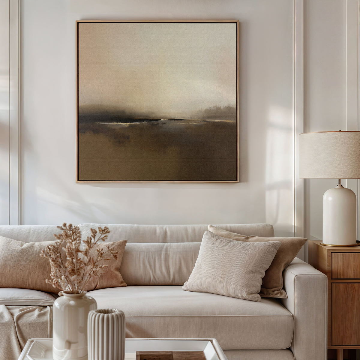 A modern living room featuring a light beige sofa, neutral pillows, wooden side table with white lamp, dried flowers in a vase, Nordic home decor, and the Foggy Beach Expanse Canvas Art - Square as a stunning focal point.