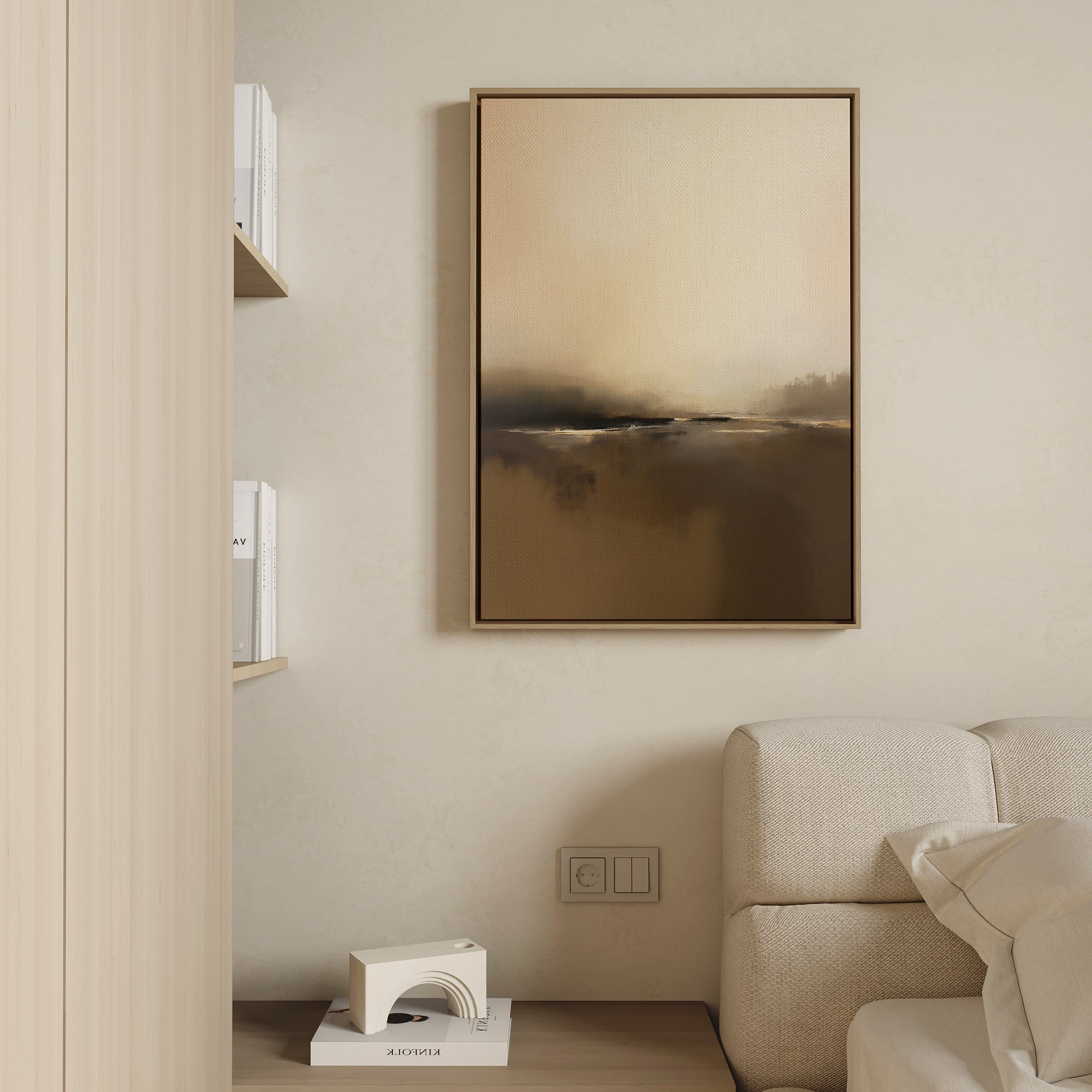 A minimalistic living room corner features a beige couch and built-in wooden shelves with white books, highlighted by the Foggy Beach Expanse Canvas Art - Vertical in a brown frame above a decorative white object on the light wall.