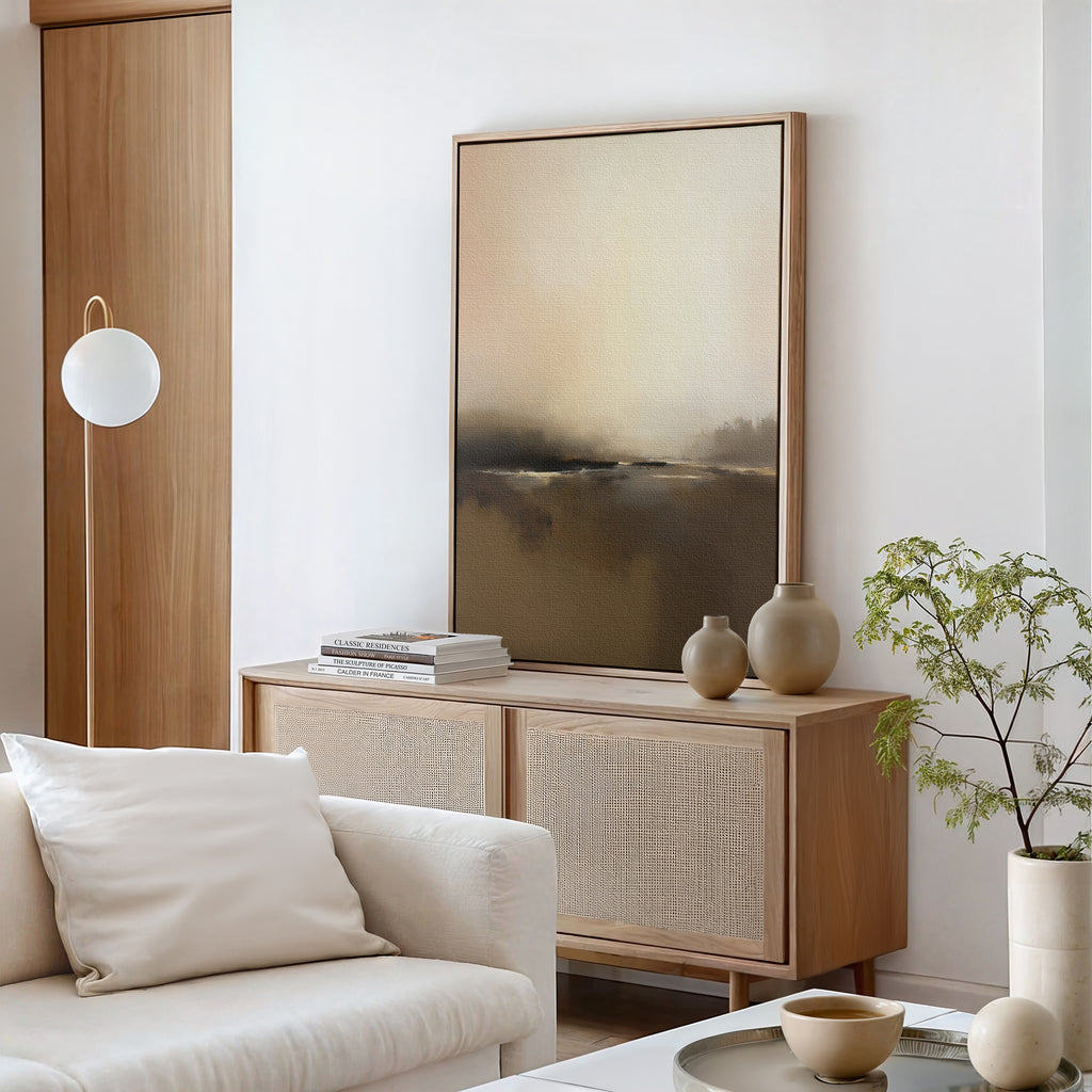 A minimalist living room features a light wood cabinet, neutral vases, stacked books, a large Foggy Beach Expanse Canvas Art - Vertical above the white sofa with a cushion, a globe floor lamp, and a small potted plant for tranquillity.