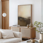 A minimalist living room features a light wood cabinet, neutral vases, stacked books, a large Foggy Beach Expanse Canvas Art - Vertical above the white sofa with a cushion, a globe floor lamp, and a small potted plant for tranquillity.