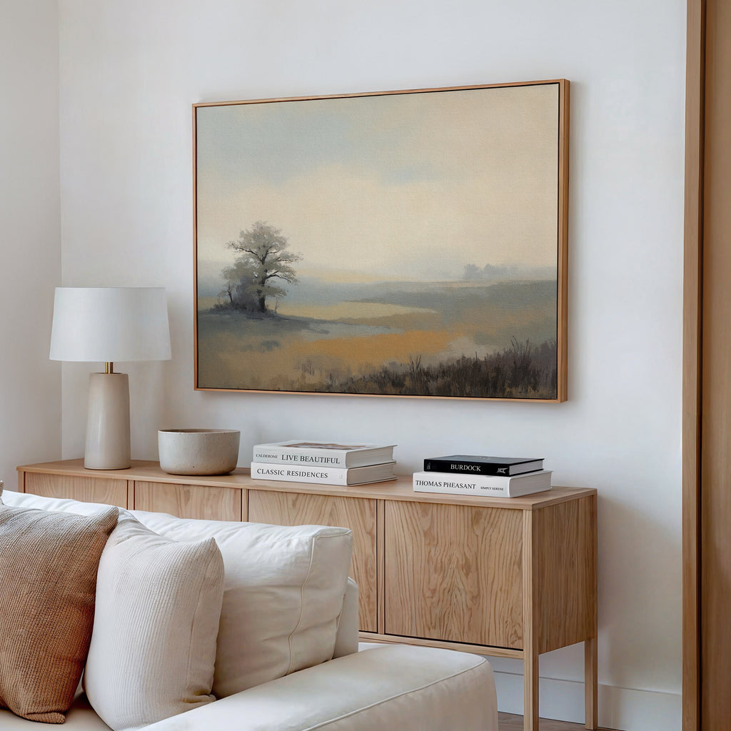 A serene living room features a wooden sideboard with stacked books, a white lamp, and the Foggy Solitude Canvas Art - Horizontal depicting a misty tree scene hanging above a beige sofa.