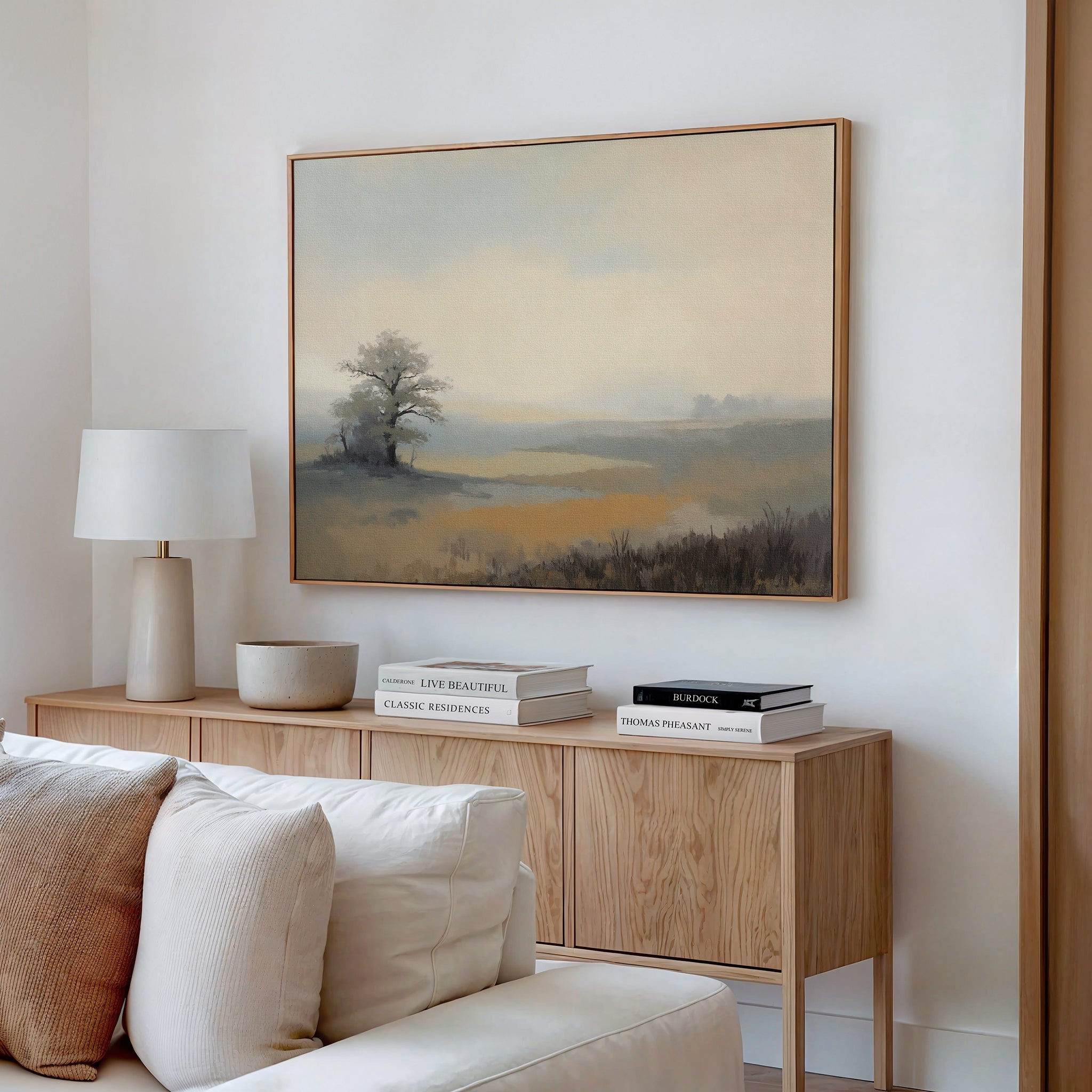 A serene living room features a wooden sideboard with stacked books, a white lamp, and the Foggy Solitude Canvas Art - Horizontal depicting a misty tree scene hanging above a beige sofa.