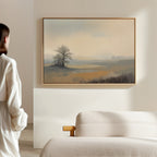 A woman in a white robe admires the "Foggy Solitude Canvas Art - Horizontal," featuring a misty landscape with a tree and fields, displayed in a frame above her light couch in a minimal, modern living room.