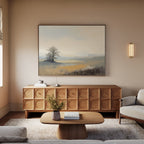 A minimalist living room features a wooden sideboard, coffee table, light sofa, and the Foggy Solitude Canvas Art - Horizontal above the sideboard. A small lamp, books, and a vase with flowers complete this serene space.